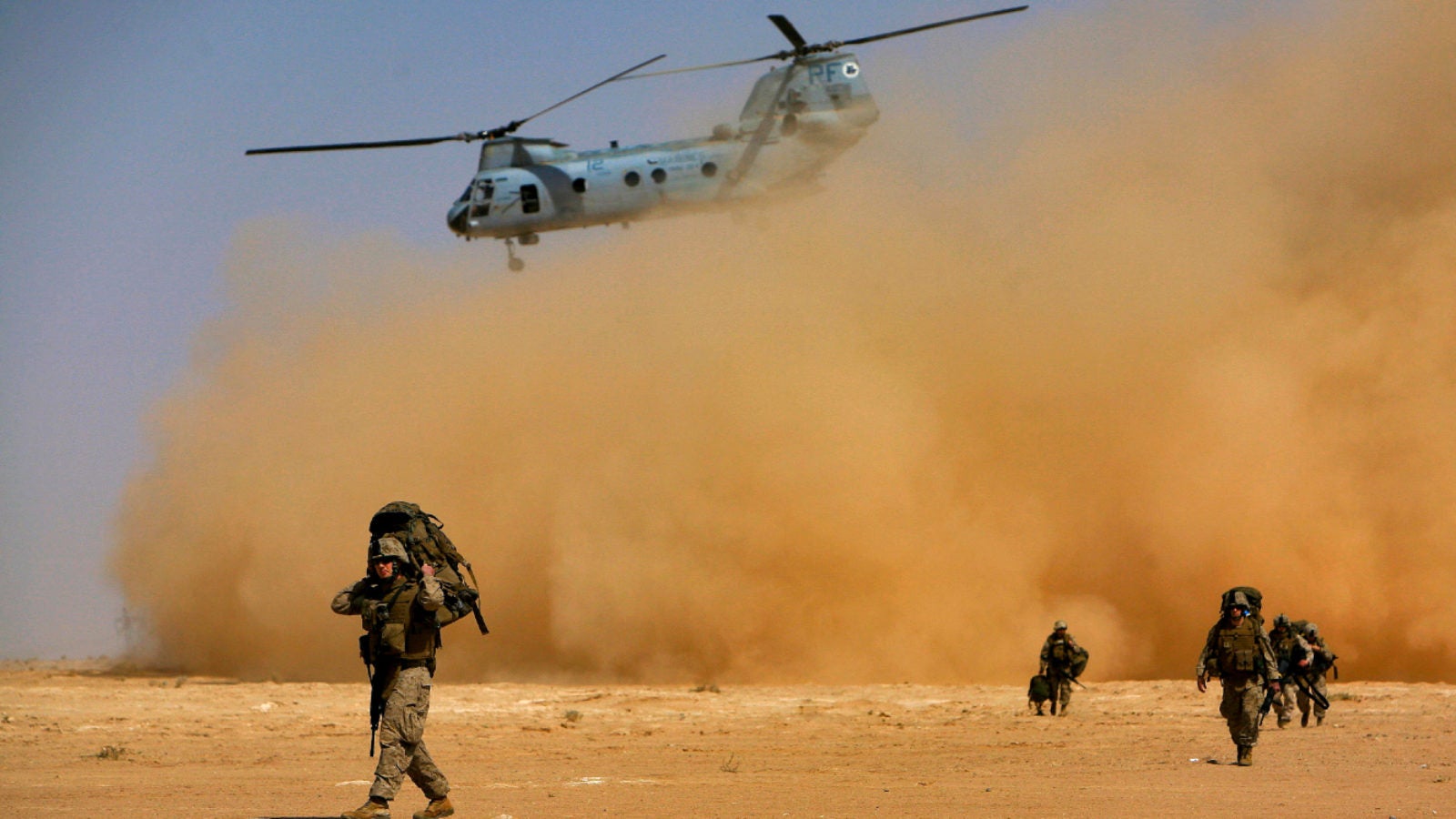 Soldiers walk in a desert underneath a departing Chinook helicopter.