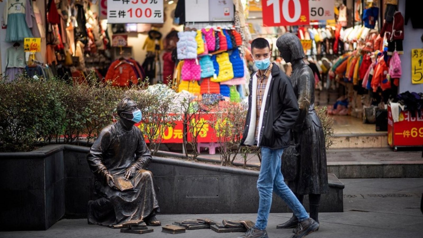 A person wearing a surgical mask walks by a storefront and a statue also wearing a mask.