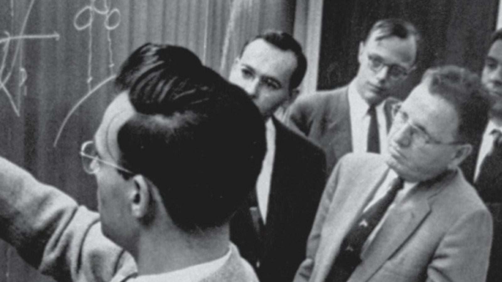 A monochrome photo of men in suits looking at graphs on a blackboard.