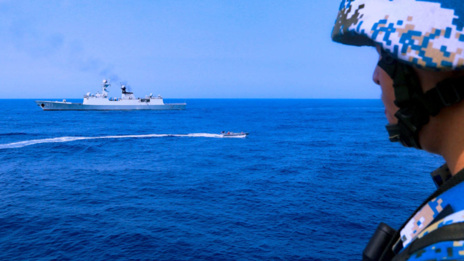 A sailor in blue camouflage looks out to the sea, where a naval vessel is on the horizon.