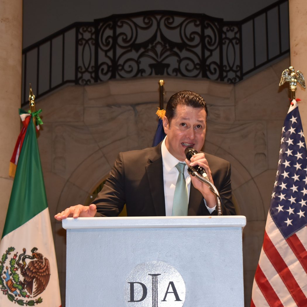 Christian Clay speaks at a podium in front of the US and Mexican flags