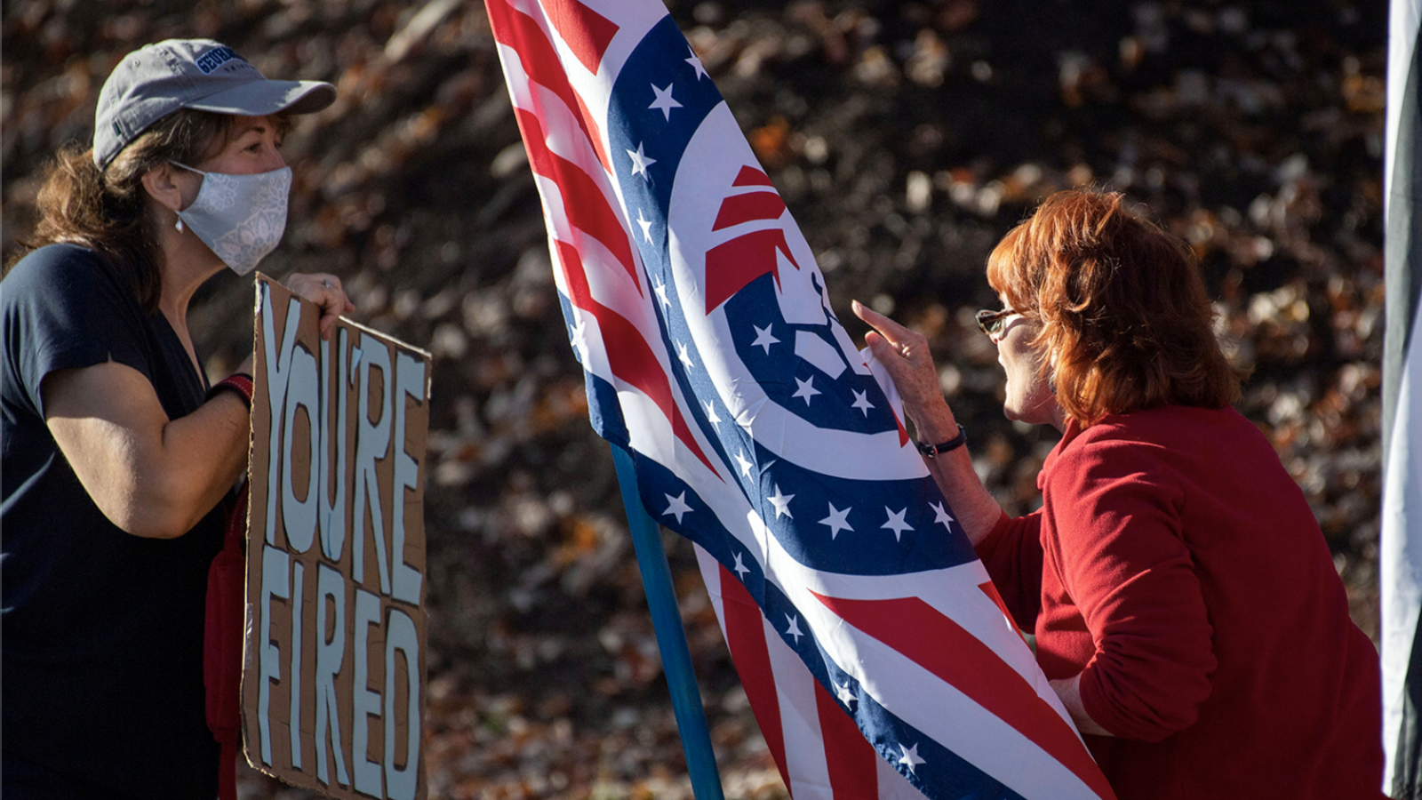 Two women, on holding a &quot;You&#039;re Fired&quot; sign and the other holding an unidentified flag argue with each other.