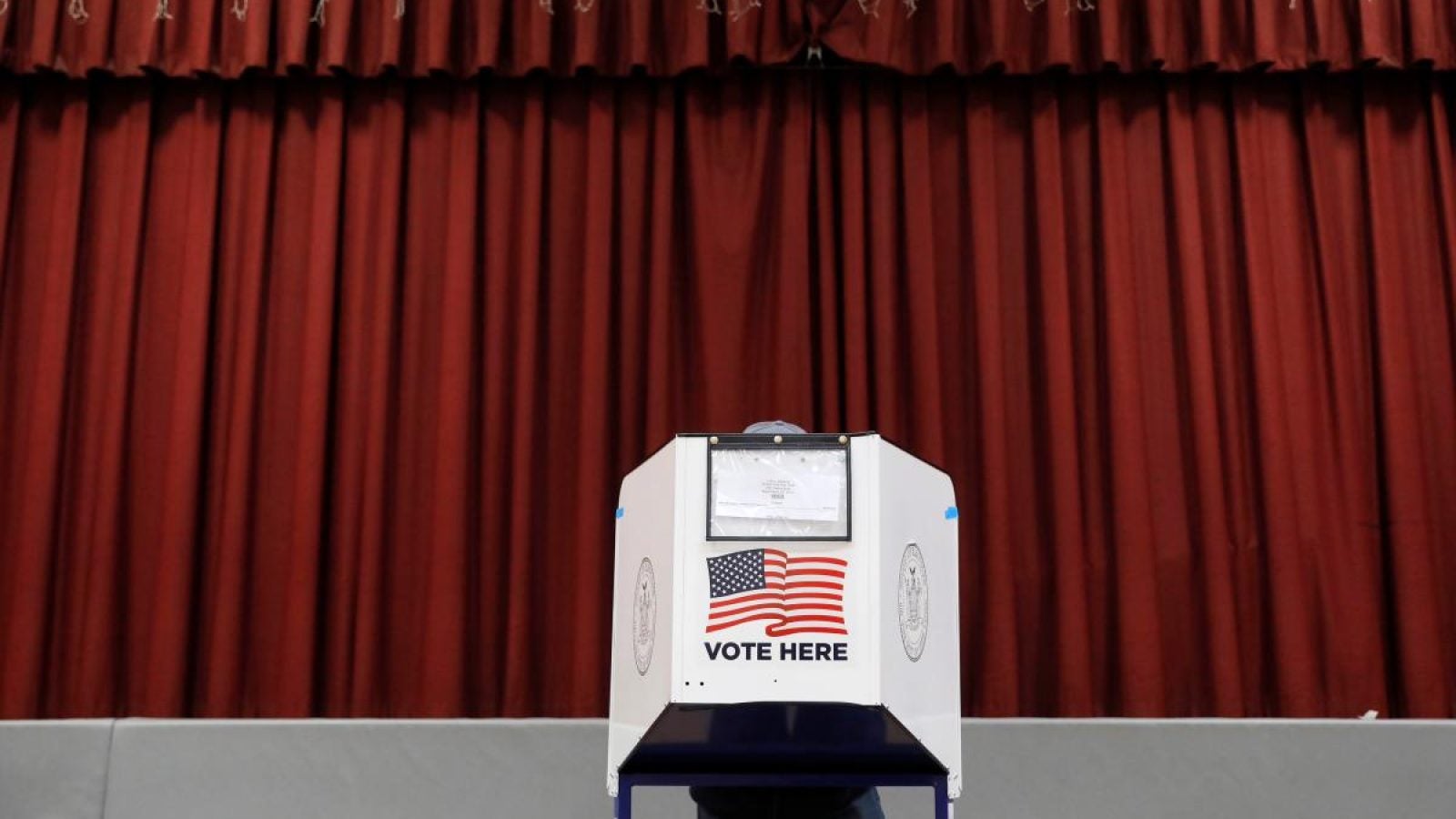 A person is hidden behind a polling booth that says &quot;Vote Here&quot; with an American flag