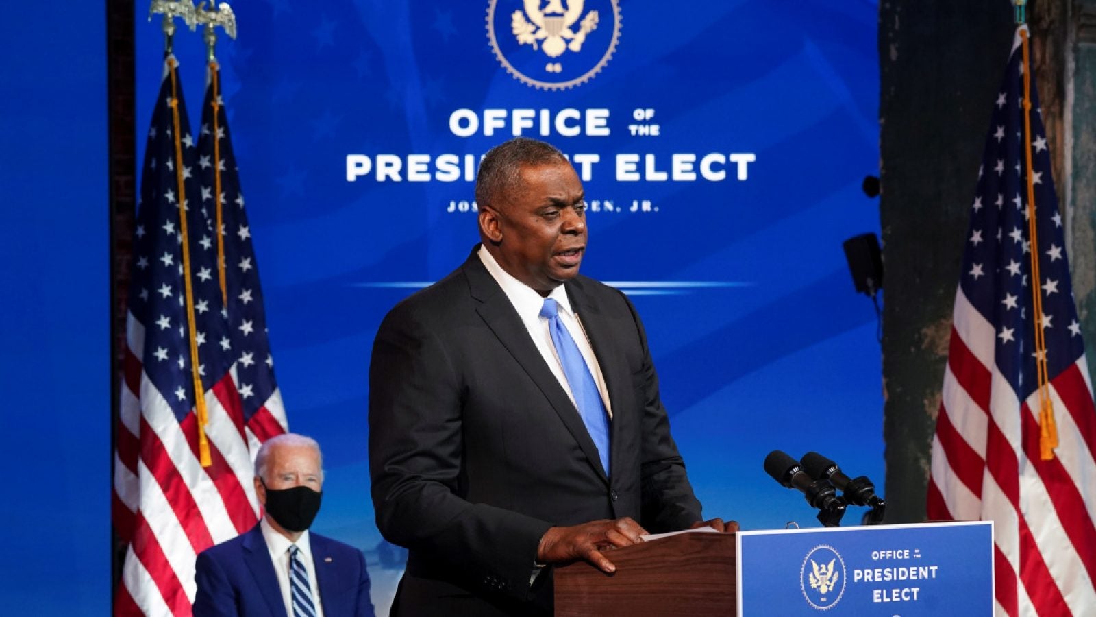 General Lloyd Austin speaks at a podium; President-elect Joe Biden and two American flags are in the background