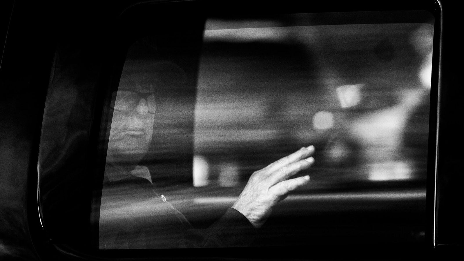 A black and white photo of Donald Trump waving from inside of a car.