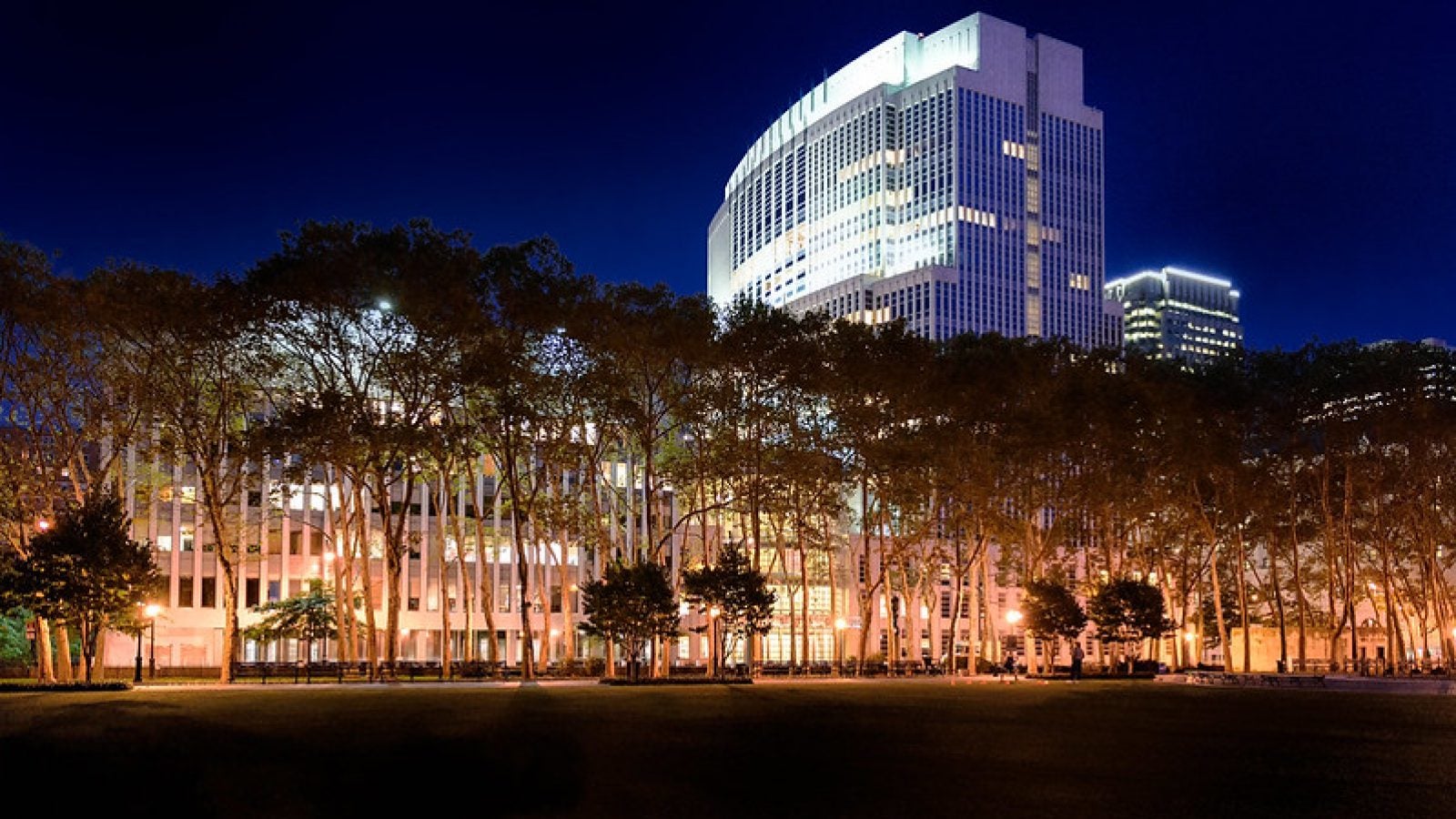 A federal courthouse is lit up at night behind a lower row of buildings and trees.