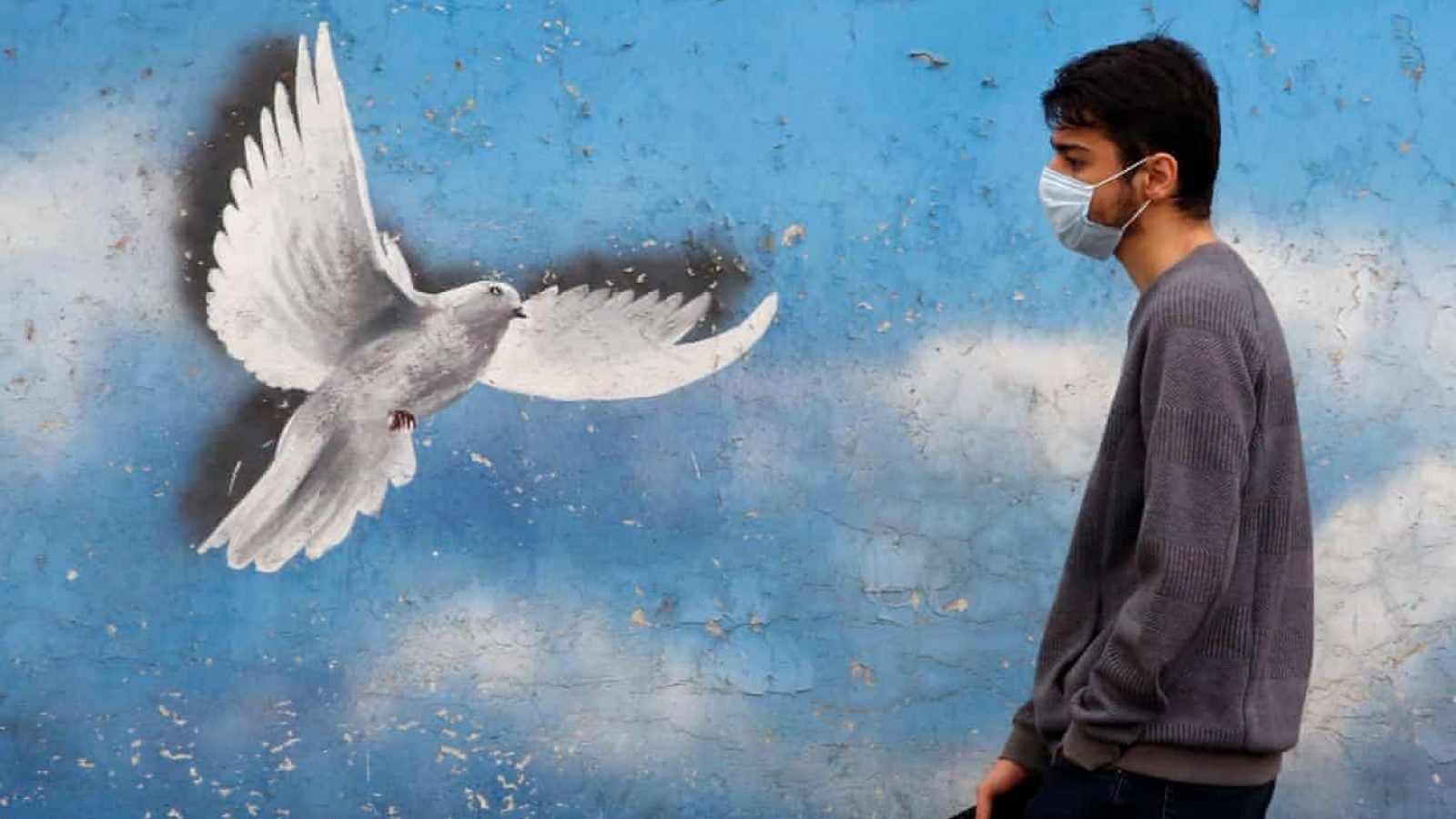 A man in a medical mask walks by a mural of a flying dove against a sky background