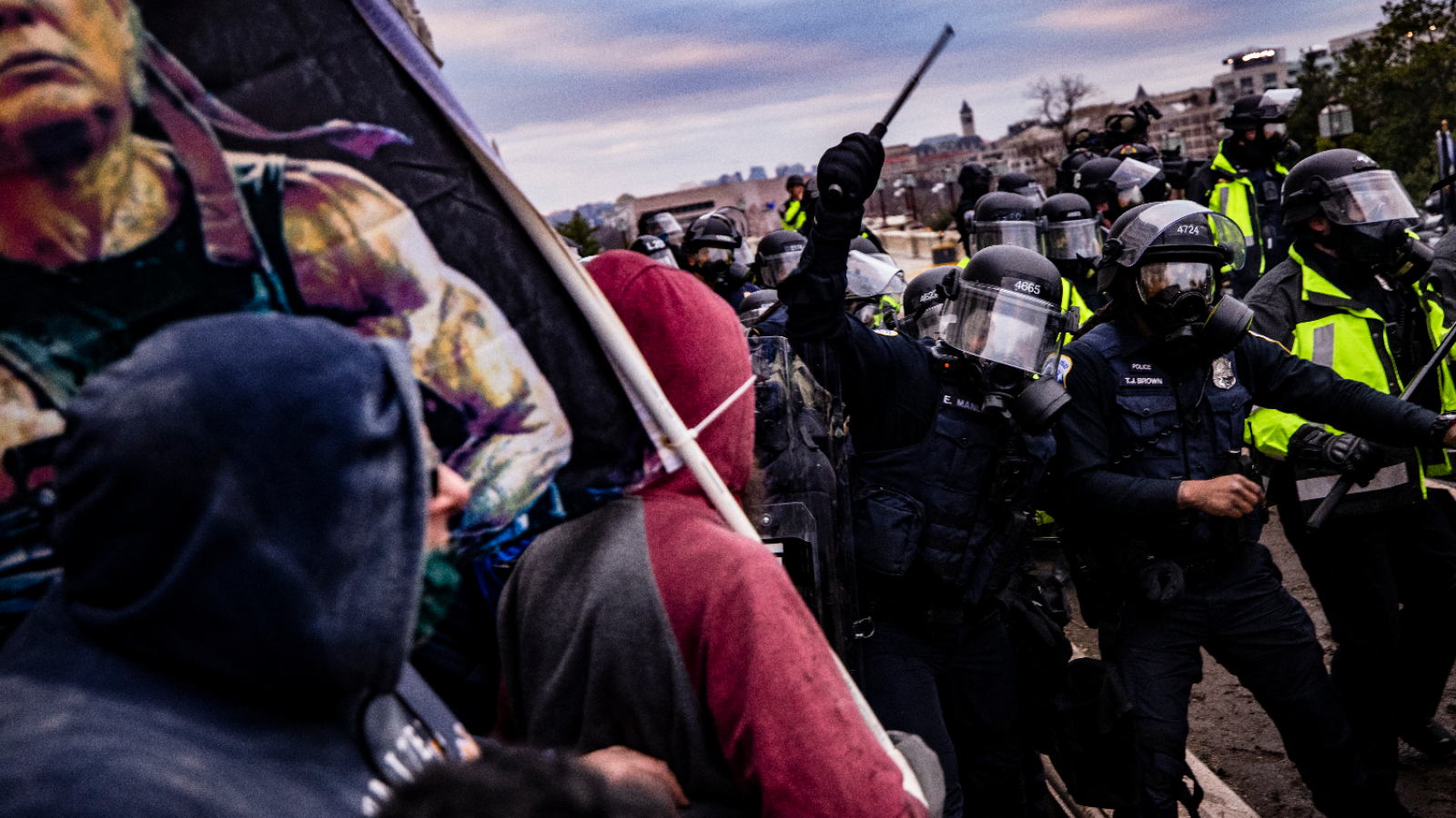 Police officers in helmets and wielding batons attempt to push back a crowd carrying Trump flags