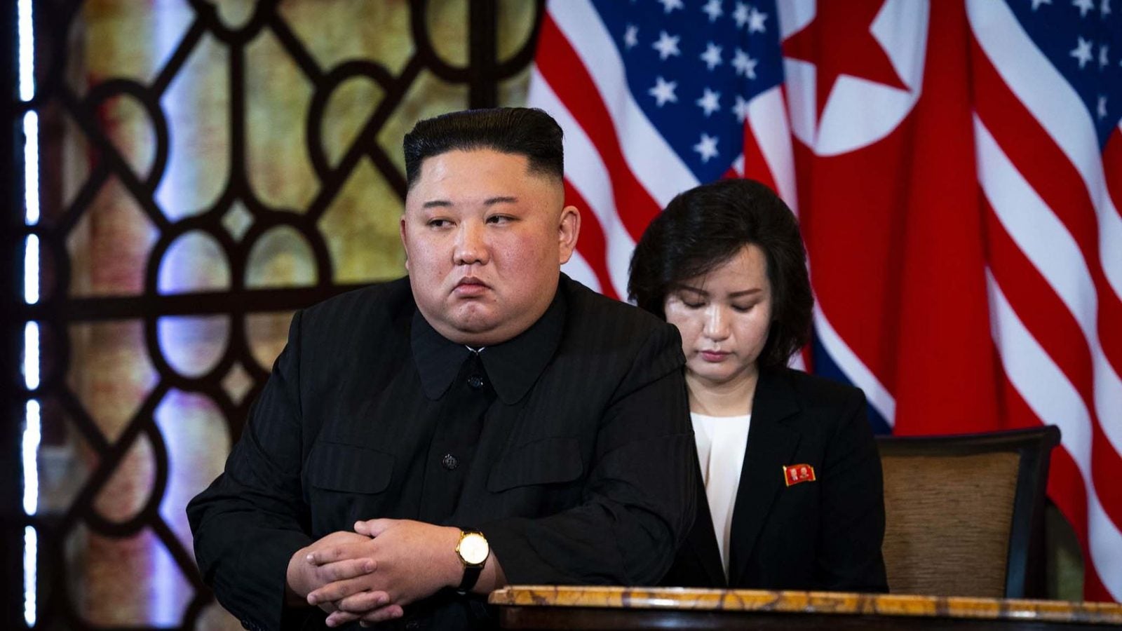 Kim Jon-un sits at the front of a table with a woman behind him and North Korean and American flags in the background.