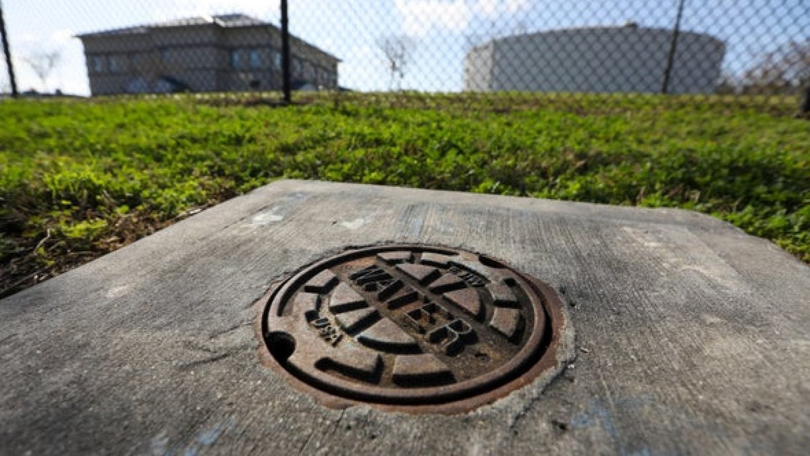 A photo of a manhole that says &quot;Water&quot; is in the foreground, with grass and buildings in the background.