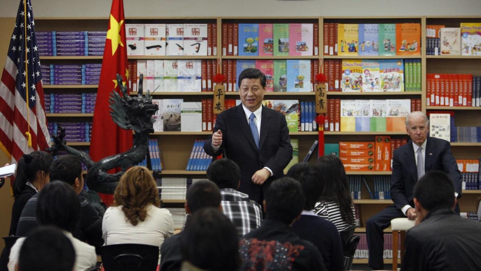 Xi Jinping points at a classroom of students while a sitting Joe Biden looks on in the background