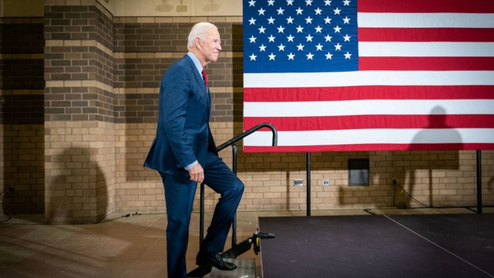 Joe Biden walkings up a small set of steps onto a stage with a large American flag in the background