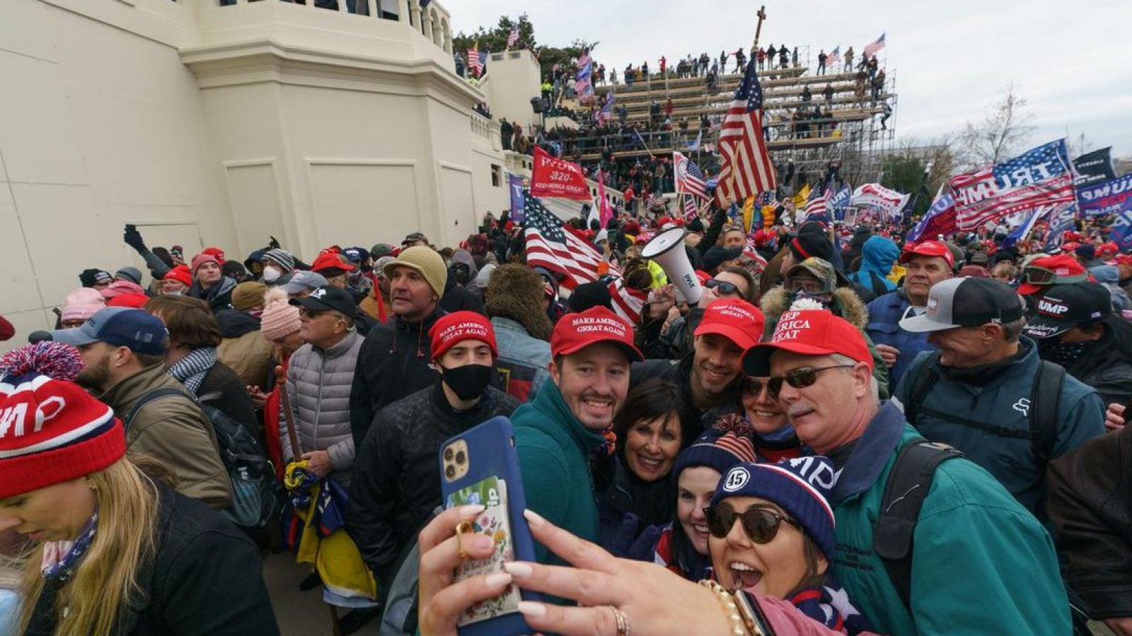 A large crowd of rioters, many wearing red MAGA hats, crowd and take selfies in front of the Capitol steps.