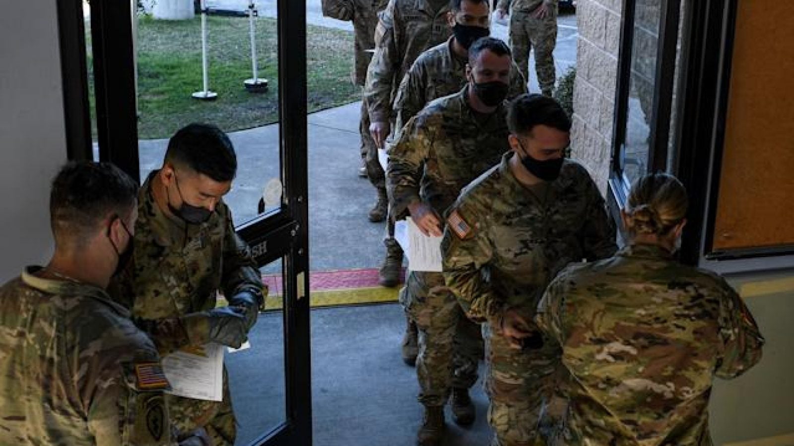 Members of the U.S. military in green camouflage uniforms line up through a door