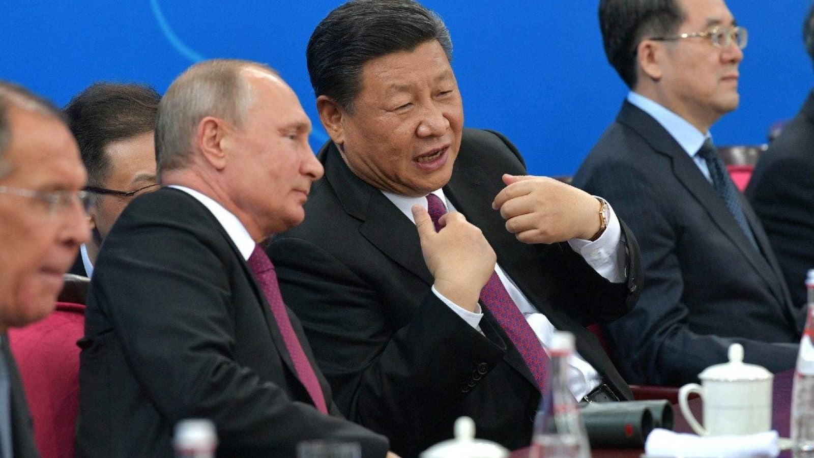 Xi Jinping speaks to Vladimir putin as they sit among other men at a table