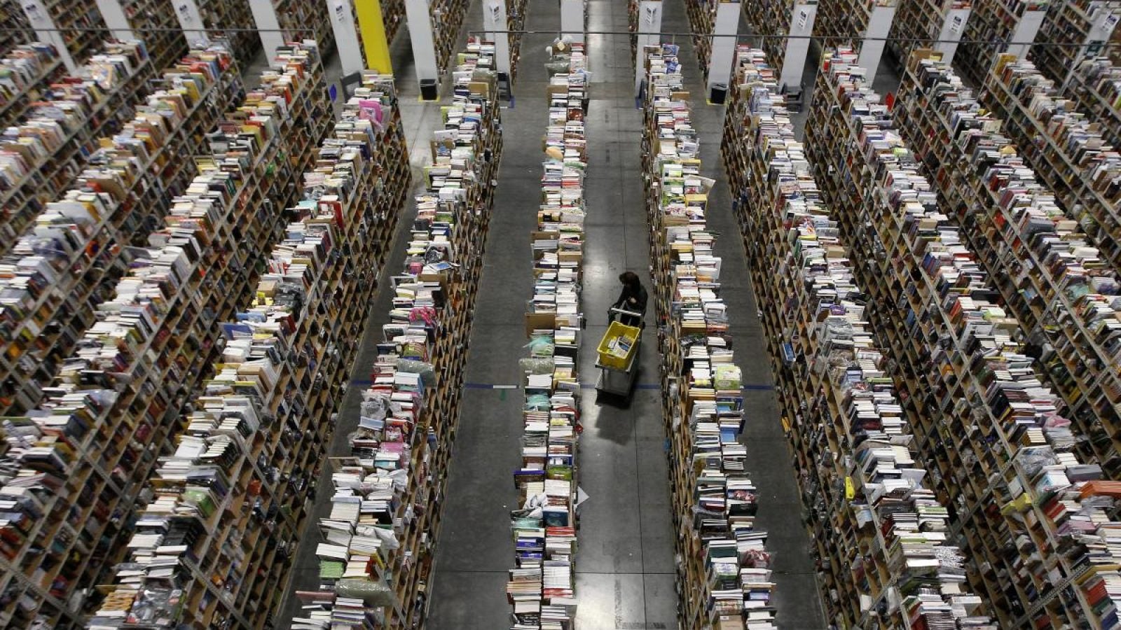 A person walks through the center aisle of large shelves filled with items in a warehouse