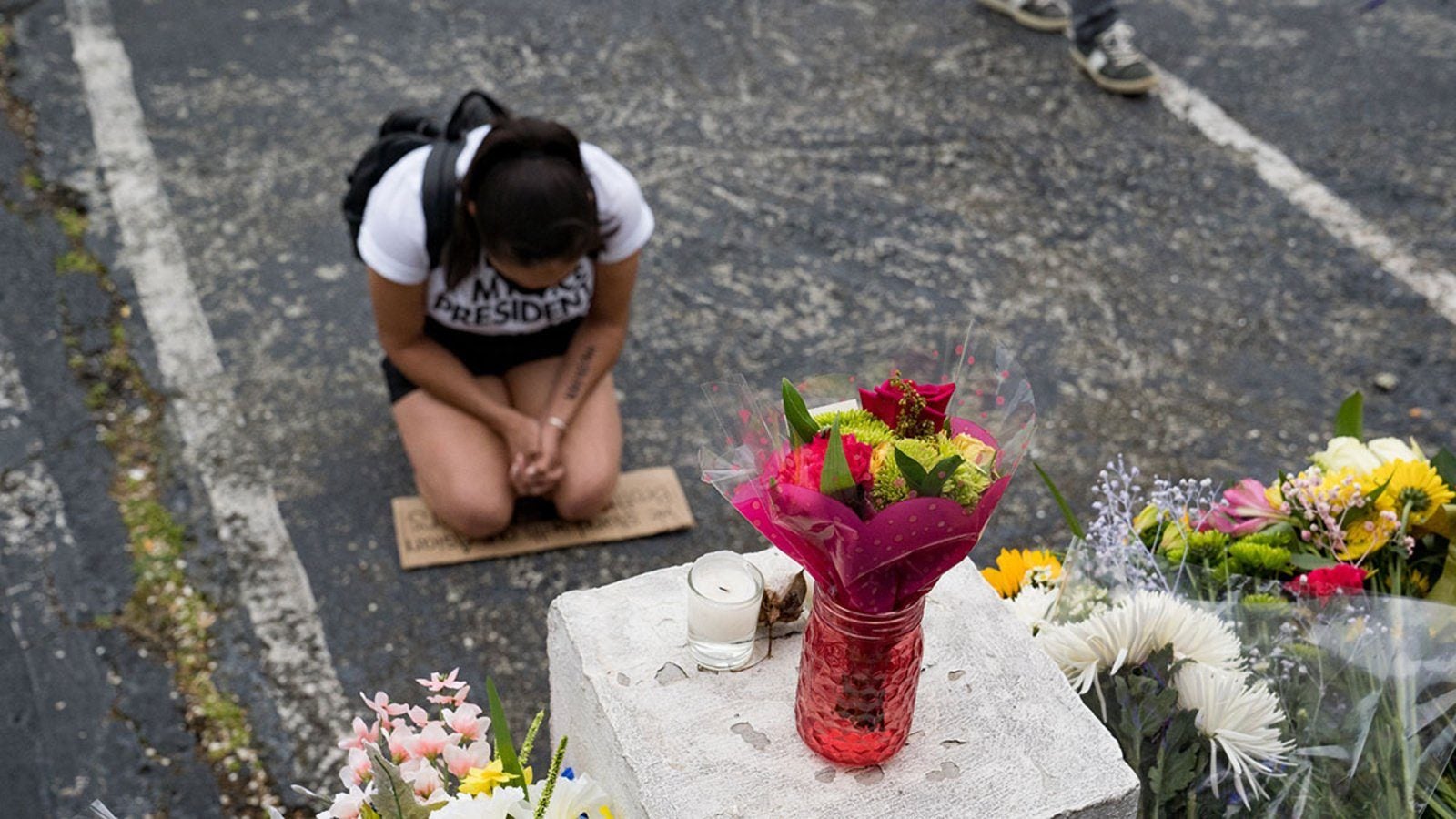 A woman kneels on a concrete parking lot in front of bouquets of flowers