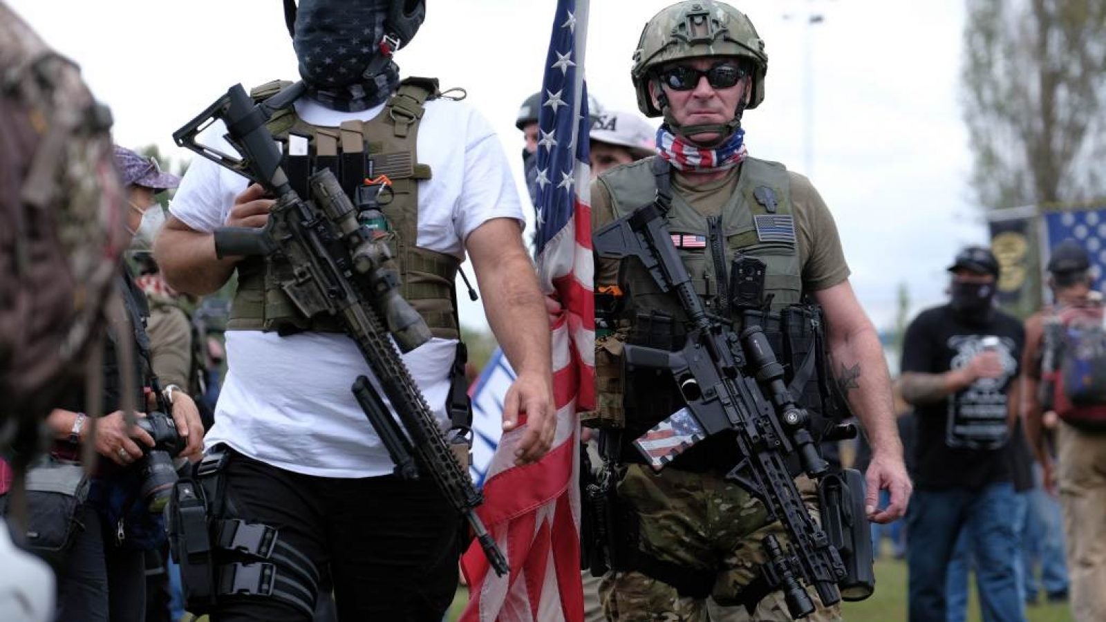 Men wearing bulletproof vests carry rifles and a large American flag