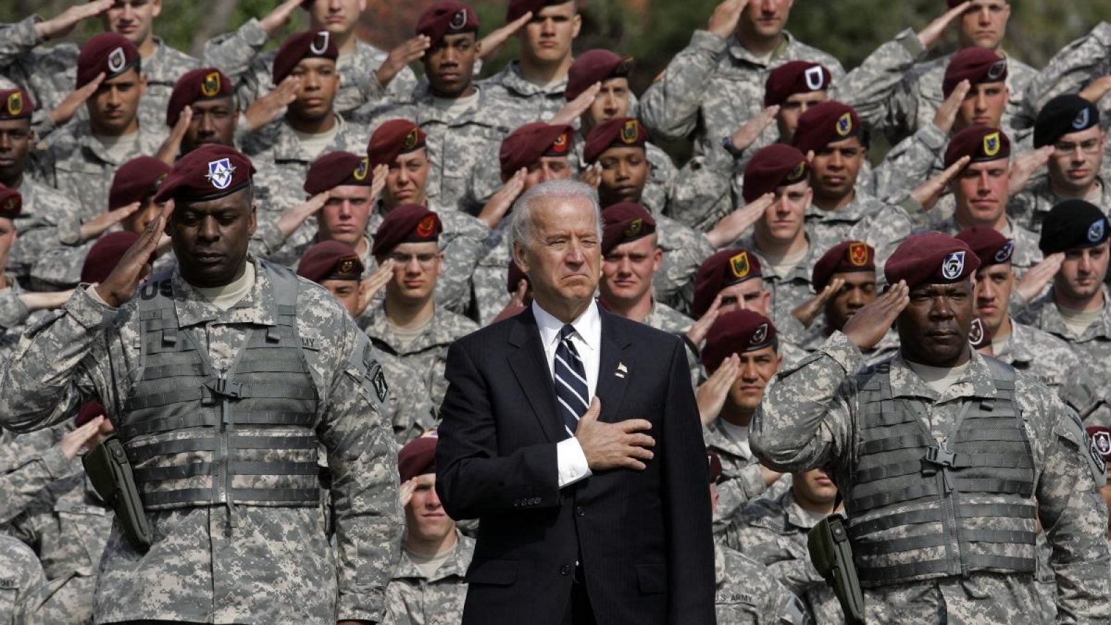 Joe Biden in a suit puts his hand over his heart and and Lloyd Austin in a green camouflage uniform salutes in the foreground, with soldiers in green camouflage saluting behind them