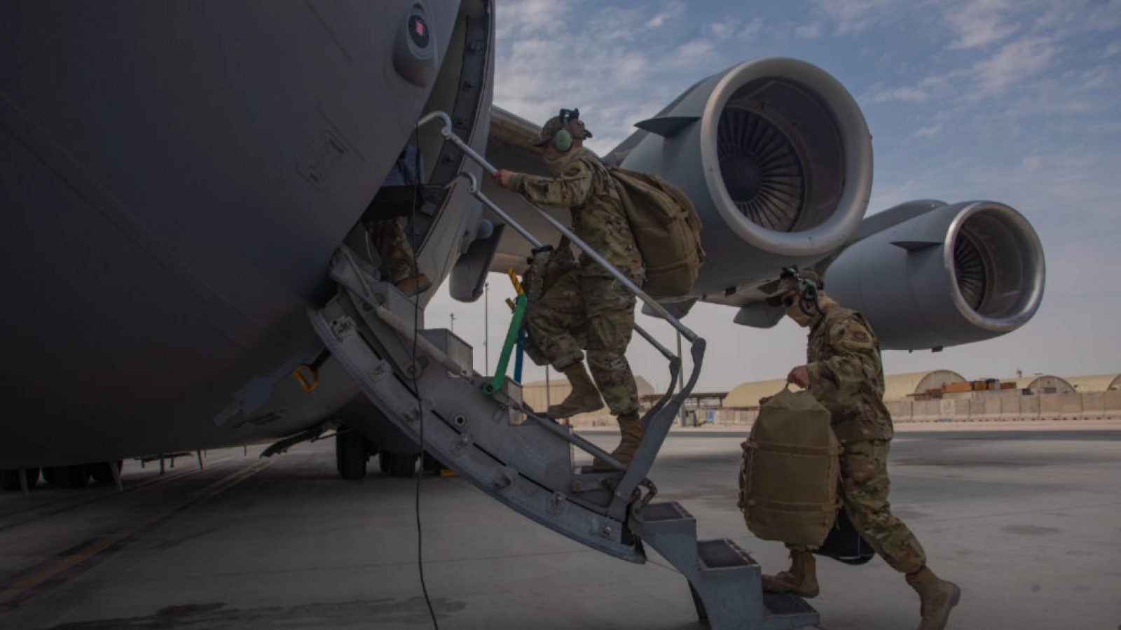 People in camouflage uniforms carry bags up a short staircase into a military plane