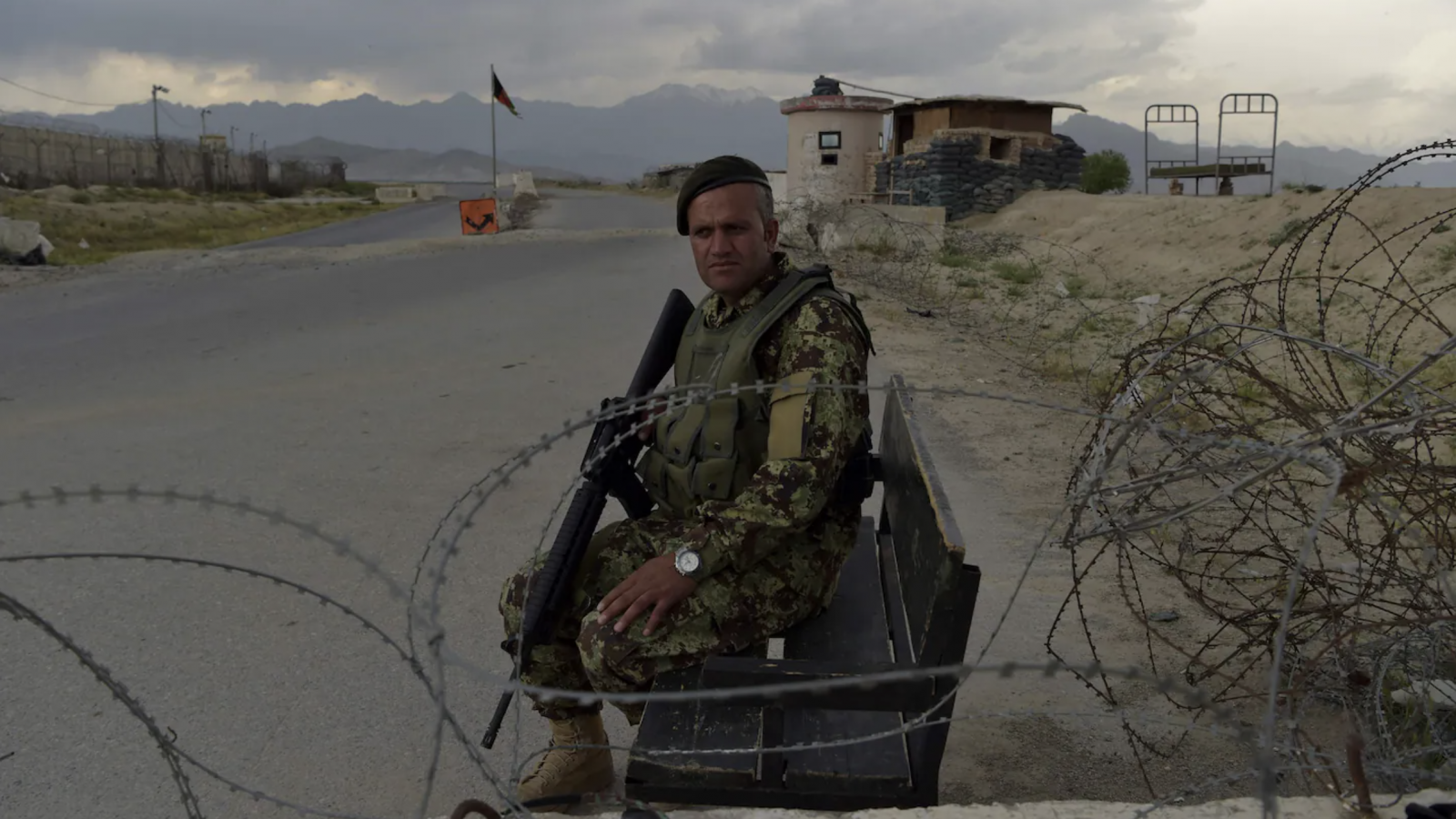 An Afghan soldier in green camouflage holds a black rifle as he sits by a road behind barbed wire