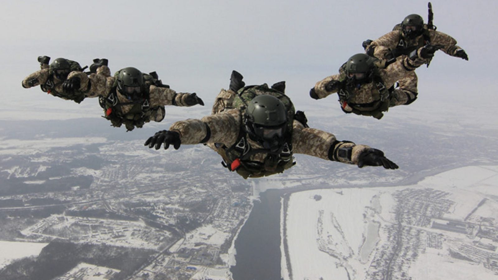 People in military uniforms are posed mid-jump from a plane over a wintry landscape