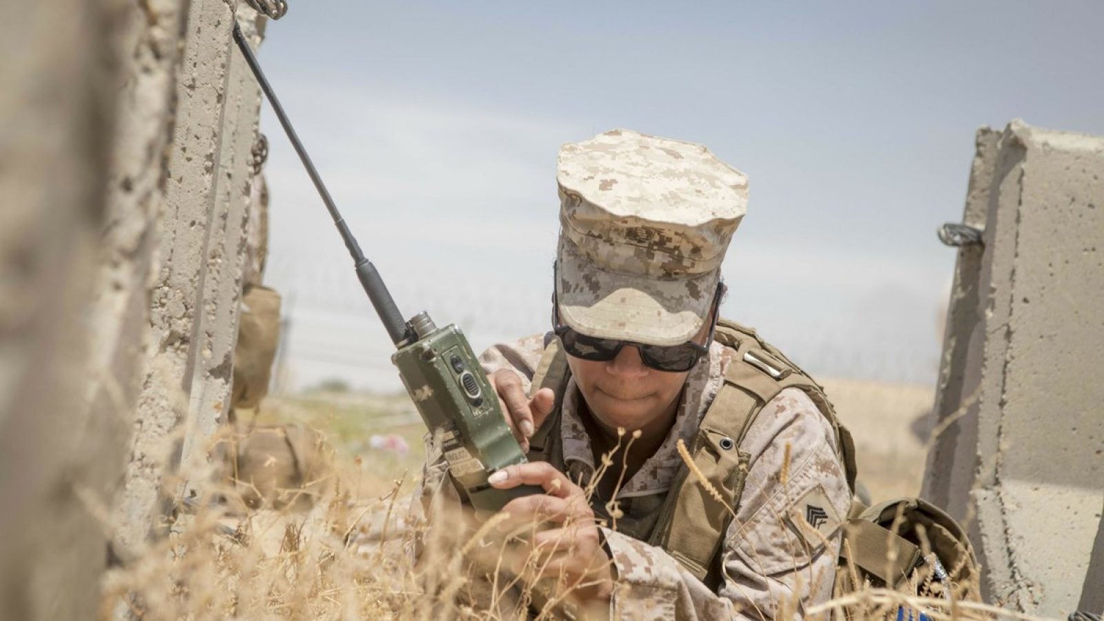 A woman in a beige military camouflage uniform and sunglasses crouches behind concrete barriers while holding a radio