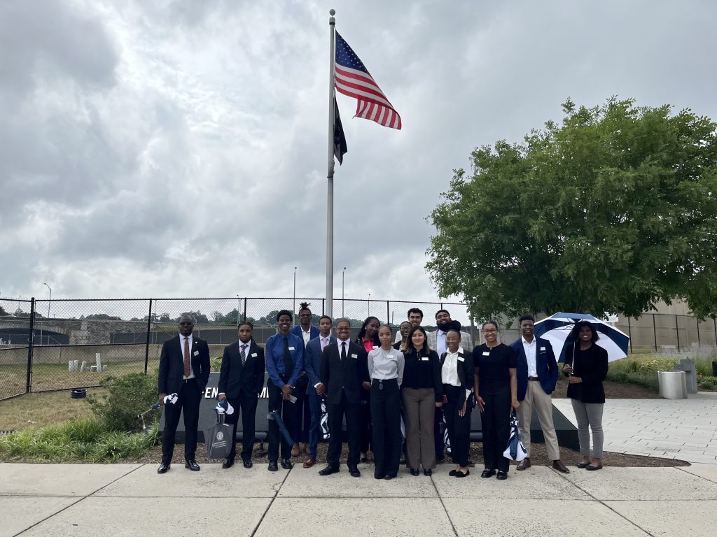 The Sumer Institute students stand outside, in front of a flagpole flying the American flag. The sky is cloudy and there is a tree to the right of the students gathered for the photo.